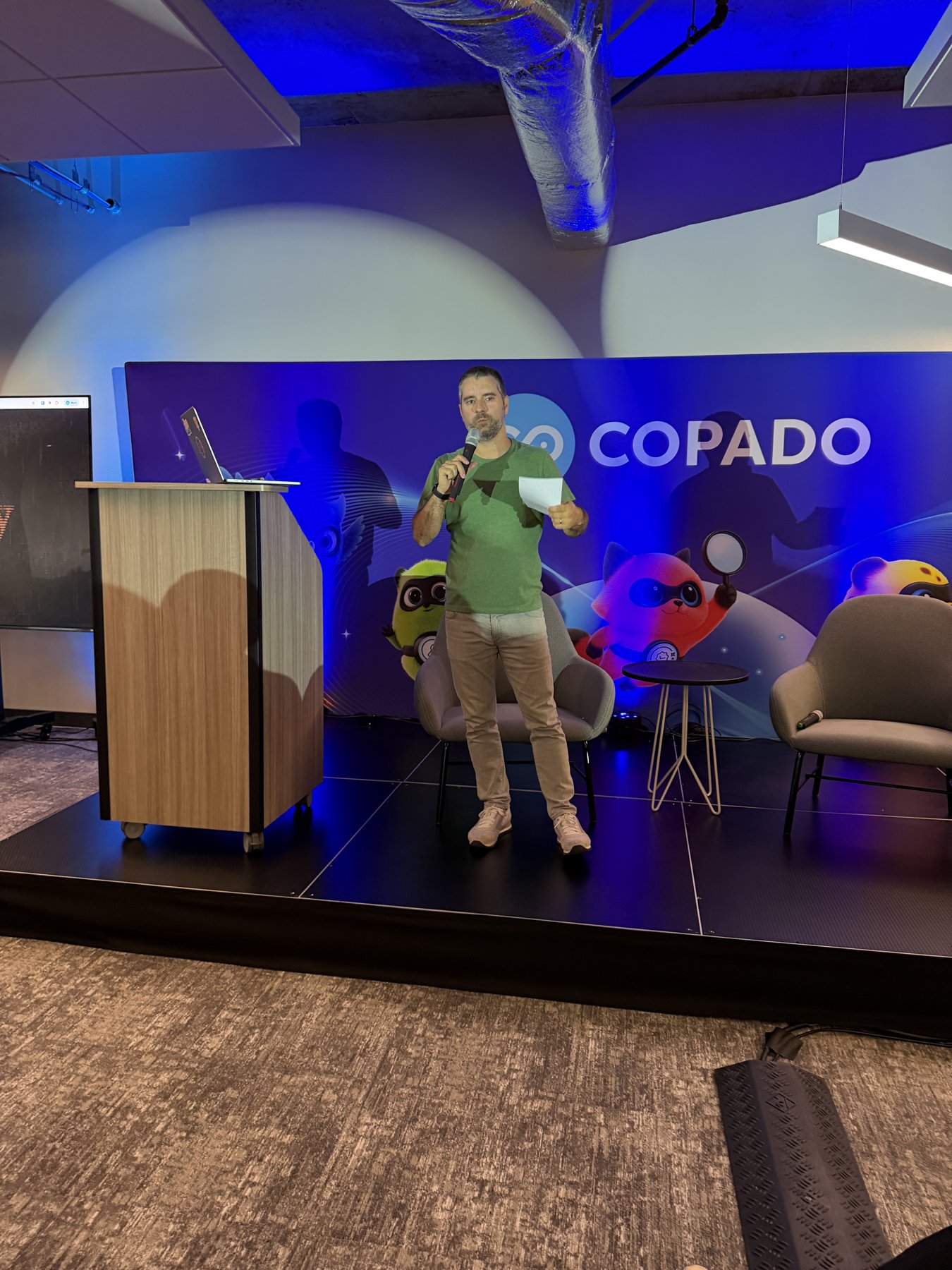 A speaker holding a microphone on stage in front of a Copado backdrop at AI Friday #6.